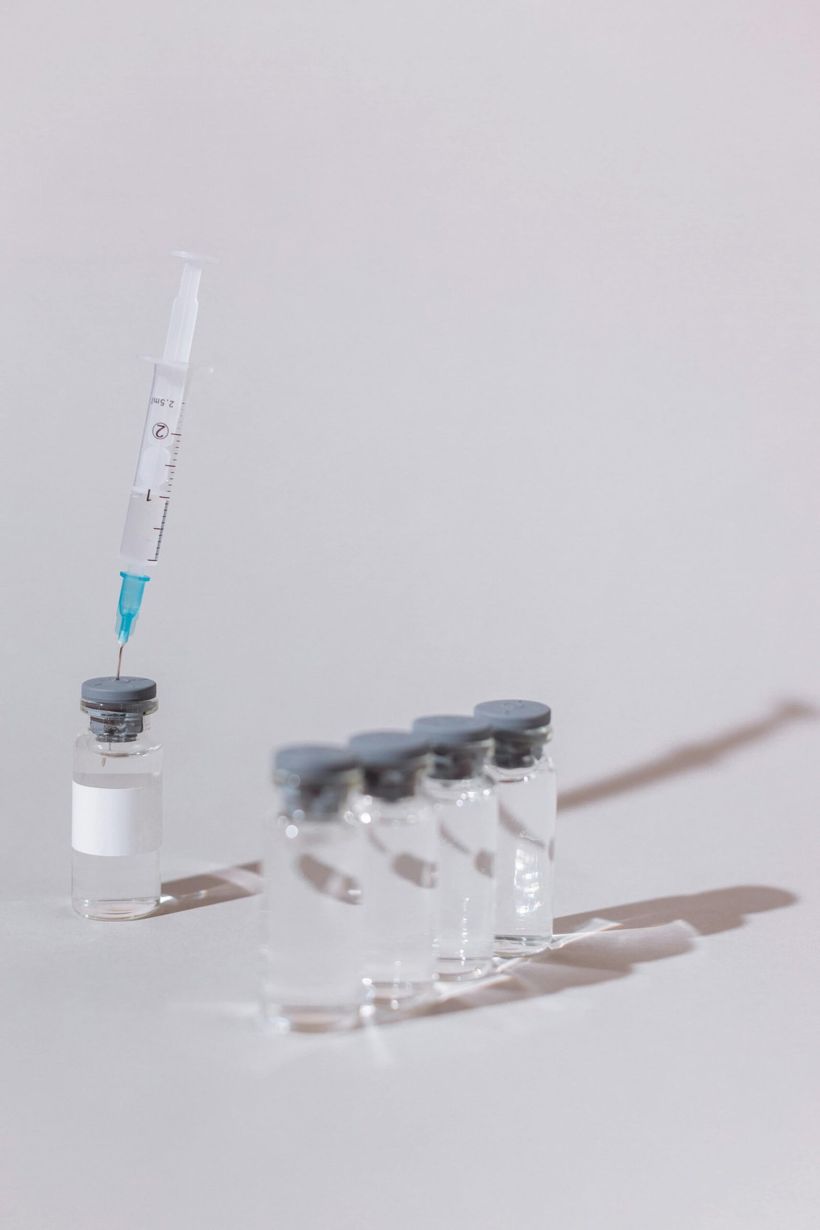 A syringe with a clear substance is placed into a glass vaccine vial on a white surface.