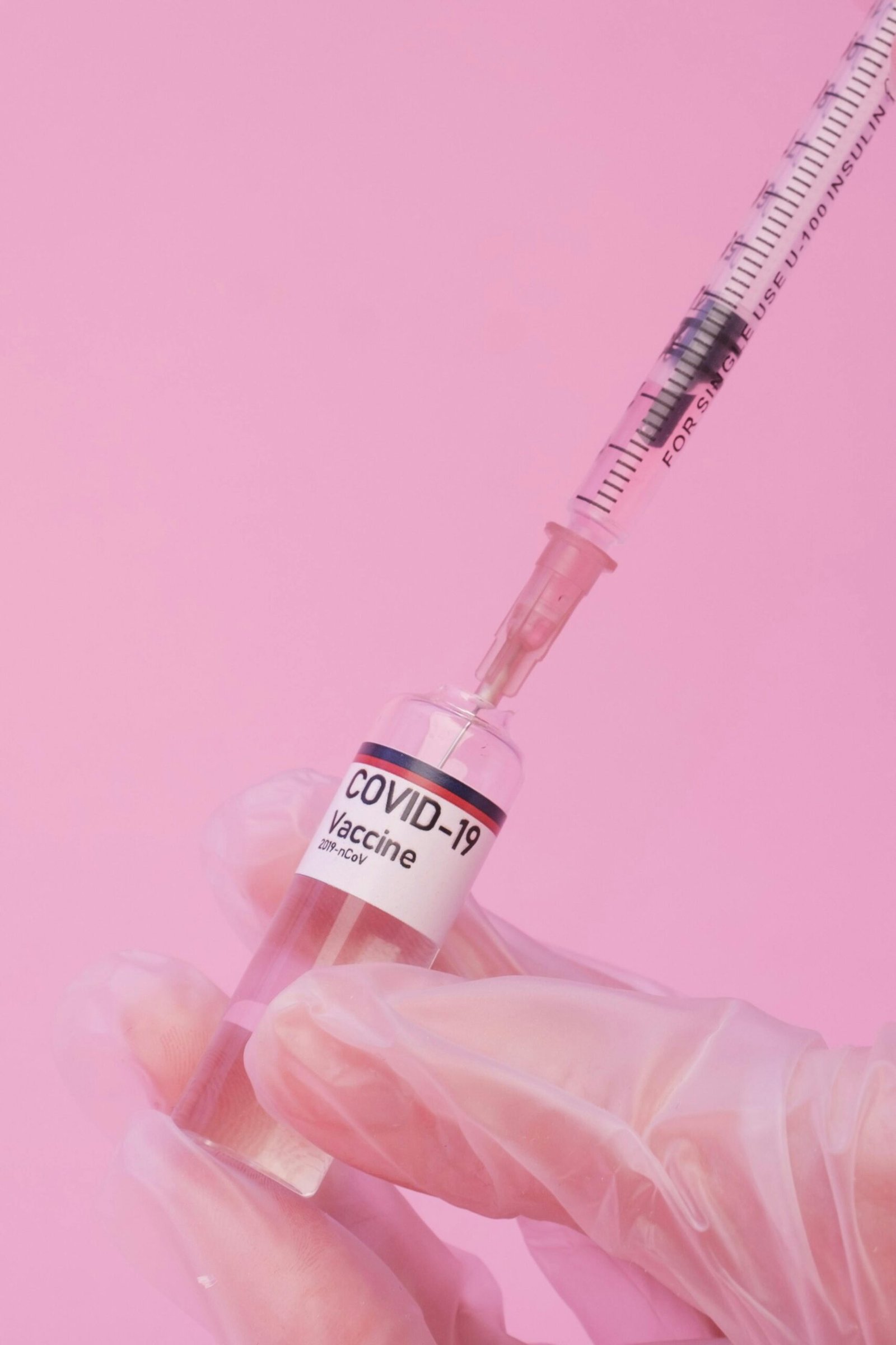 Crop anonymous female doctor in medical gloves filling syringe with COVID 19 vaccine from ampoule against pink background