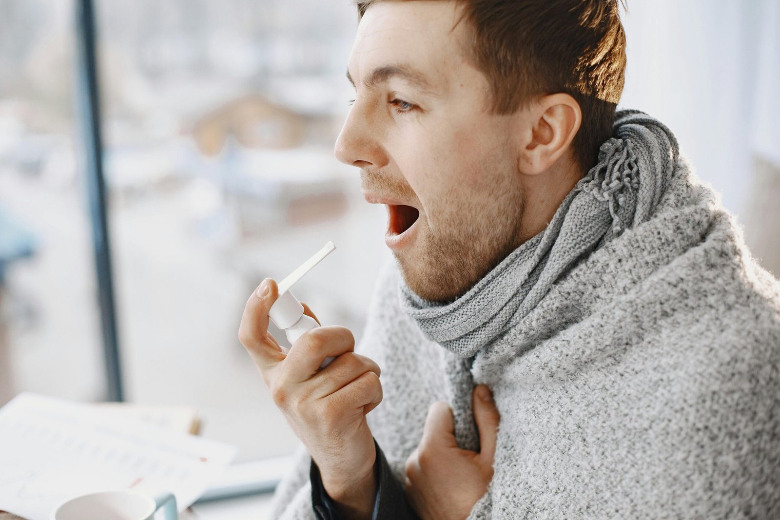 An adult man uses a throat spray, wrapped in a blanket, appearing unwell indoors.